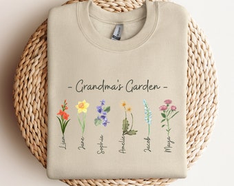 May include: A beige sweatshirt with the text "Grandma's Garden" printed on the front.  The sweatshirt features a row of six watercolor flowers with names written below each flower: Liam, Jane, Sophia, Amelie, Jacob, and Maya.