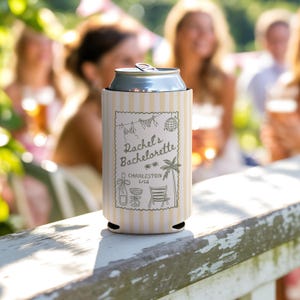 May include: A white and yellow striped can cooler with the text "Rachel's Bachelorette Charleston 2026" and various illustrations. The cooler is holding a silver can and is resting on a white wooden surface.