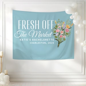 May include: A light blue bachelorette party banner with the text "Fresh Off The Market" and a bouquet of pink, yellow, and white flowers. The banner also includes the text "Katie's Bachelorette Charleston, 2025".