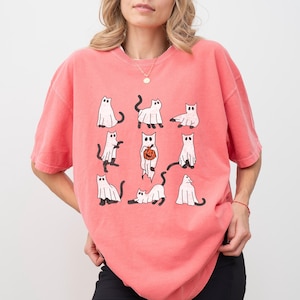 Puede incluir: Una camiseta rosa con un gr&aacute;fico de nueve gatos blancos vestidos de fantasmas. Los gatos est&aacute;n en varias poses, incluyendo sosteniendo una calabaza.