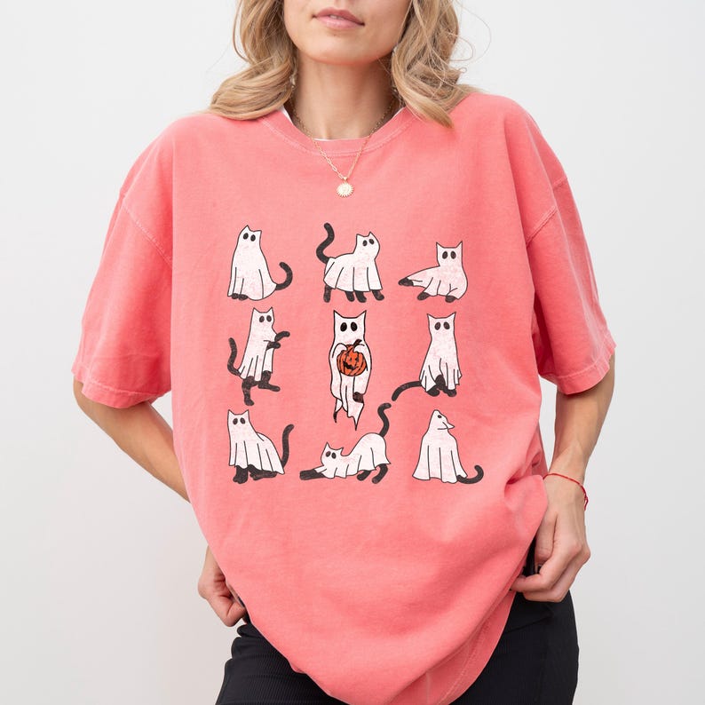 Puede incluir: Una camiseta rosa con un gr&aacute;fico de nueve gatos blancos vestidos de fantasmas. Los gatos est&aacute;n en varias poses, incluyendo sosteniendo una calabaza.