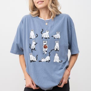 Puede incluir: Una camiseta azul con un estampado gr&aacute;fico de nueve gatos blancos vestidos de fantasmas. Algunos de los gatos sostienen calabazas.