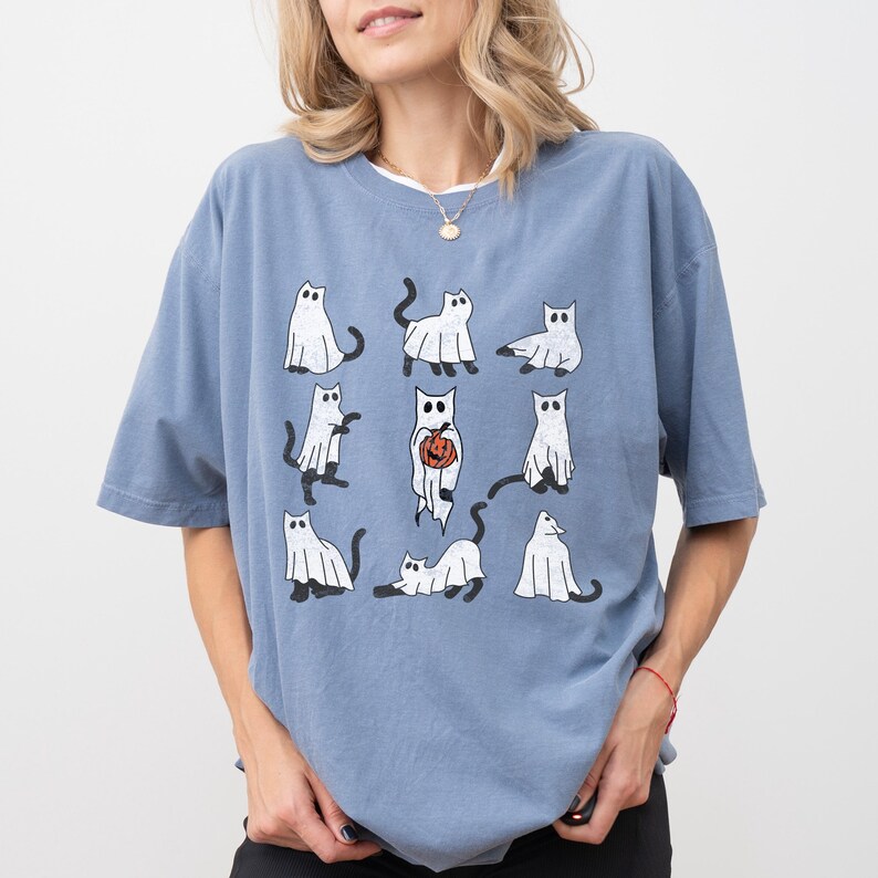 Puede incluir: Una camiseta azul con un estampado gr&aacute;fico de nueve gatos blancos vestidos de fantasmas. Algunos de los gatos sostienen calabazas.