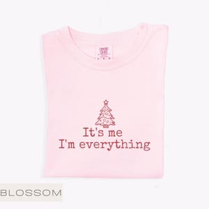May include: A pale pink t-shirt featuring the text "It's me I'm everything" and a Christmas tree design. The shirt is folded neatly, with a tag visible at the collar. The word "BLOSSOM" is at the bottom left.