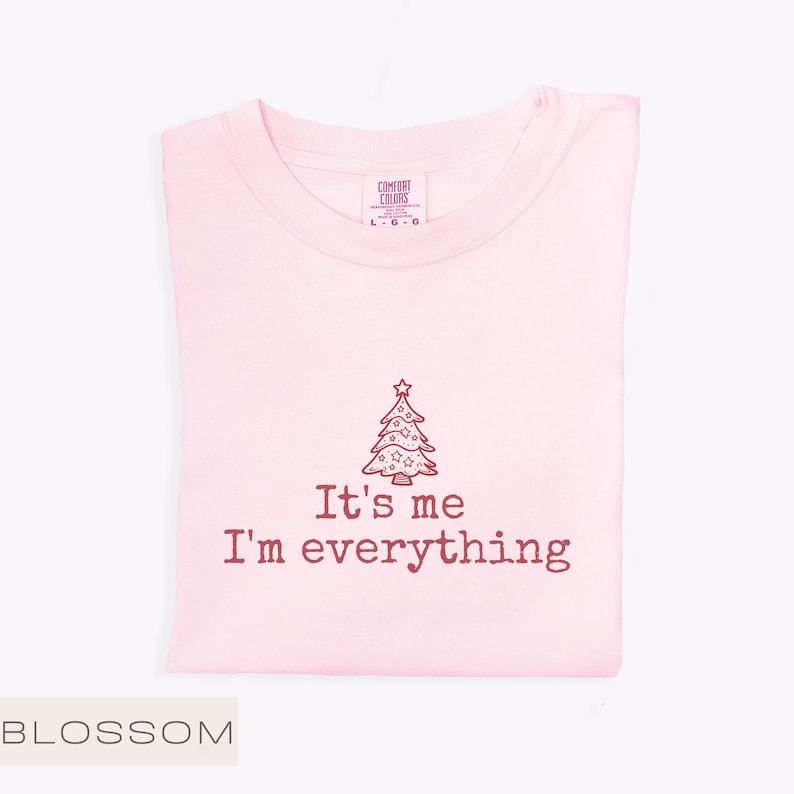 May include: A pale pink t-shirt featuring the text "It's me I'm everything" and a Christmas tree design. The shirt is folded neatly, with a tag visible at the collar. The word "BLOSSOM" is at the bottom left.