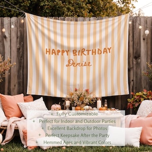 Puede incluir: Un tel&oacute;n de fondo a rayas con el texto "HAPPY BIRTHAY Denise" en letras naranjas. El tel&oacute;n de fondo cuelga frente a una valla de madera. La escena incluye almohadas, calabazas y arreglos florales, lo que sugiere una fiesta.