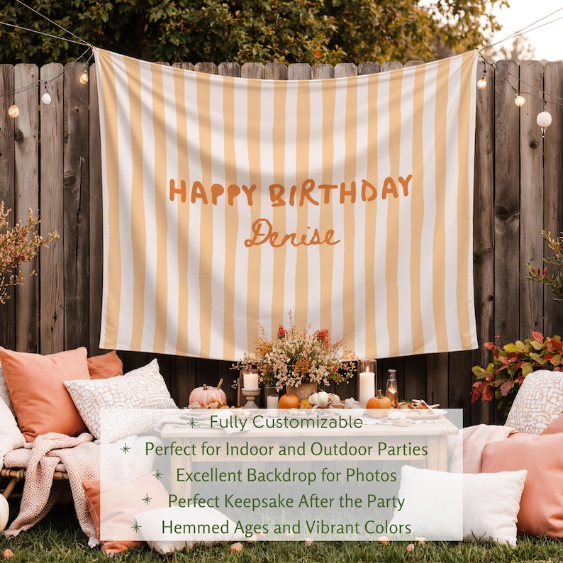 Puede incluir: Un tel&oacute;n de fondo a rayas con el texto "HAPPY BIRTHAY Denise" en letras naranjas. El tel&oacute;n de fondo cuelga frente a una valla de madera. La escena incluye almohadas, calabazas y arreglos florales, lo que sugiere una fiesta.