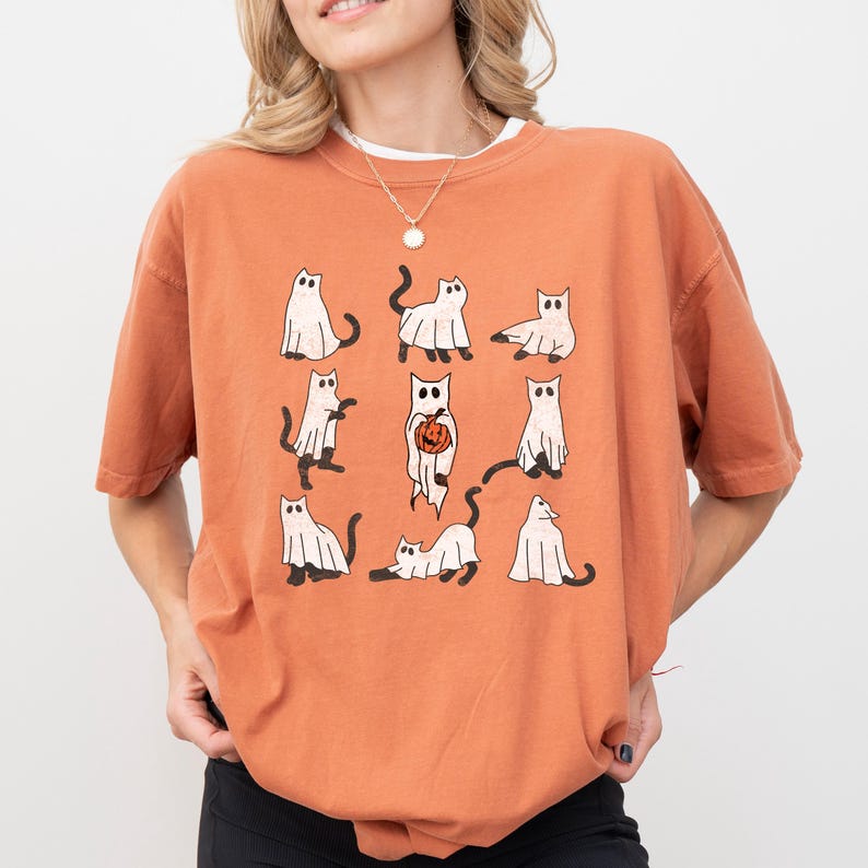 Puede incluir: Camiseta naranja con un gr&aacute;fico de gatos fantasmas blancos sosteniendo calabazas. Los gatos est&aacute;n en varias poses, incluyendo sentados, de pie y caminando.