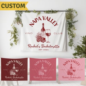 May include: A white, red, and pink fabric banner with the text "NAPA VALLEY" in a circle with the words "VINO" and "CLUB" on either side. The banner also includes a drawing of a bottle of wine, a glass of wine, and grapes. The text "Rachel's Bachelorette EST. 2024" is below the circle.