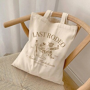 Op de afbeelding: Een witte canvas tote bag met bruine tekst die "Last Rodeo" en "Nicole's Bachelorette Nashville, TN" zegt. De tas heeft ook een afbeelding van een vrouw in een cowboyhoed die een gitaar vasthoudt en in een martini-glas zit.