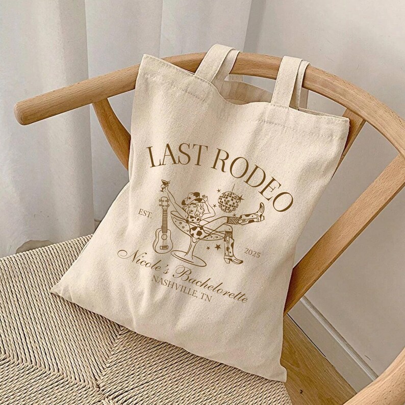 Op de afbeelding: Een witte canvas tote bag met bruine tekst die "Last Rodeo" en "Nicole's Bachelorette Nashville, TN" zegt. De tas heeft ook een afbeelding van een vrouw in een cowboyhoed die een gitaar vasthoudt en in een martini-glas zit.