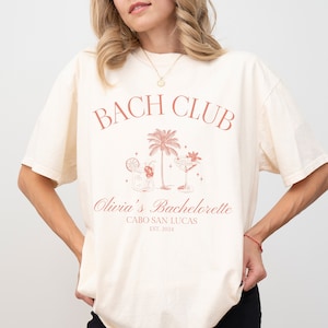 May include: A white t-shirt with a pink graphic design that says "Bach Club" with palm trees and cocktails. The text below says "Olivia's Bachelorette Cabo San Lucas Est. 2024".