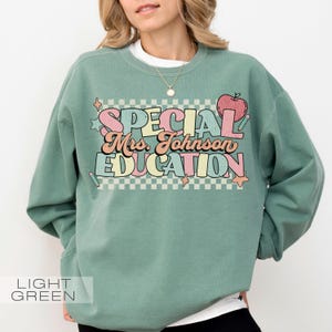 Op de afbeelding: Lichtgroene sweatshirt met de tekst "SPECIAL Mrs. Johnson EDUCATION" in een kleurrijk, retro lettertype. Het ontwerp bevat een appelafbeelding en steraccenten. De sweatshirt is een comfortabele, casual stijl.
