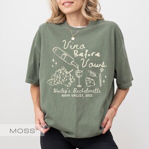 以下が含まれることがあります： モスグリーンのTシャツに白いグラフィックデザイン。デザインには、グラスに注がれるワインボトル、ブドウ、チーズ、そして「Vina Before Vows, Hailey's Bachelorette, Napa Valley, 2025」の文字が含まれています。