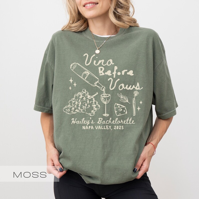 以下が含まれることがあります： モスグリーンのTシャツに白いグラフィックデザイン。デザインには、グラスに注がれるワインボトル、ブドウ、チーズ、そして「Vina Before Vows, Hailey's Bachelorette, Napa Valley, 2025」の文字が含まれています。