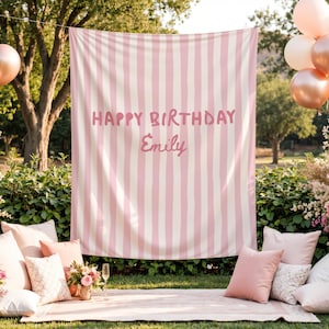 Puede incluir: Un tel&oacute;n de fondo a rayas rosas y blancas con el texto "HAPPY BIRTHDAY Emily" se muestra al aire libre. La escena incluye globos, almohadas y una manta de picnic, creando un ambiente festivo de cumplea&ntilde;os.