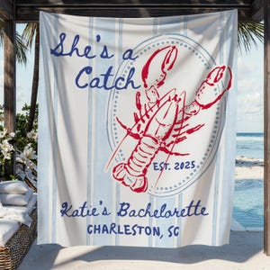 May include: A striped beach towel in blue and white, featuring the text "She's a Catch" in blue script. A red lobster graphic is centered, with "EST. 2025" below. The text "Katie's Bachelorette, Charleston, SC" is at the bottom.