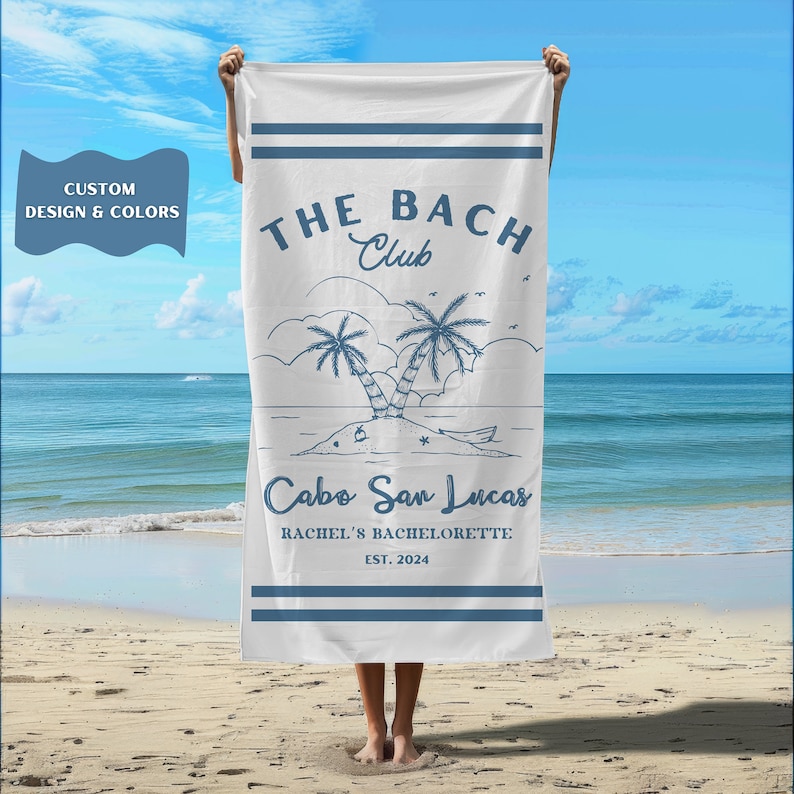 May include: White beach towel with blue text and graphics. The text reads "THE BACH Club Cabo San Lucas Rachel's Bachelorette EST. 2024".
