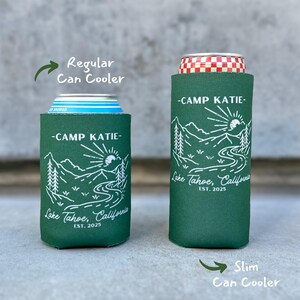 May include: Two green can coolers with a white mountain scene and the text "Camp Katie - Lake Tahoe, California - Est. 2025". The can cooler on the left is a regular size and the can cooler on the right is a slim size.