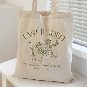 Op de afbeelding: Een witte canvas tote bag met groene tekst die "Last Rodeo" zegt en een afbeelding van een vrouw in een cowboyhoed en laarzen die in een martini-glas zit. De tekst bevat ook "Est. 2025" en "Nicole's Bachelorette Nashville, TN".
