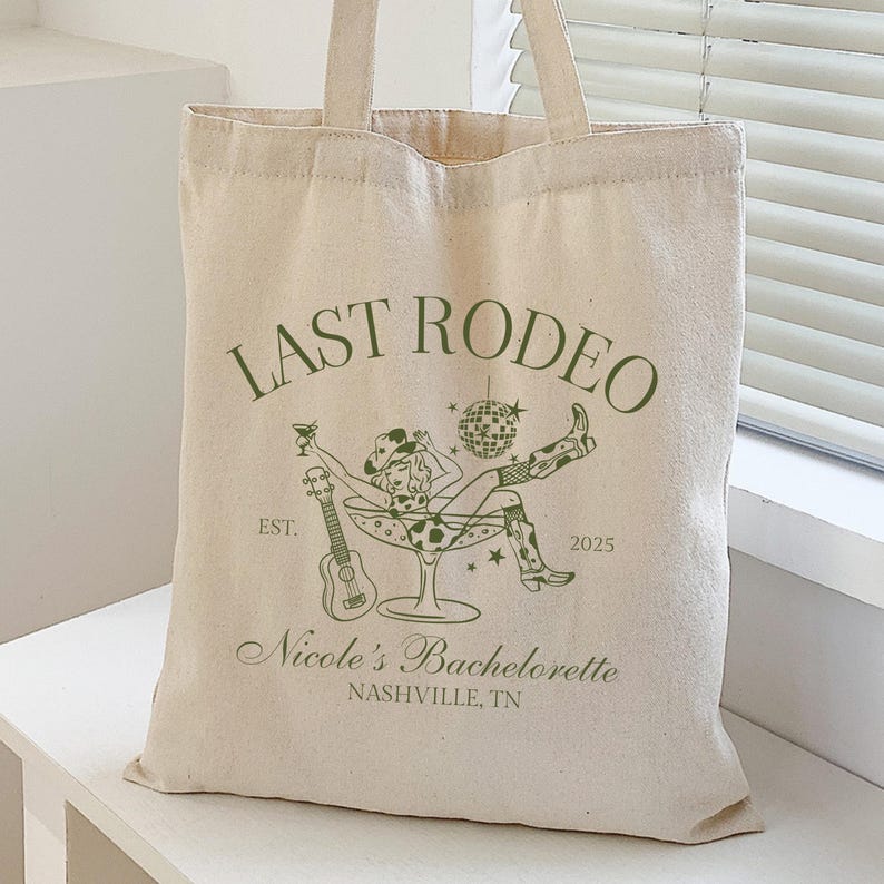 Op de afbeelding: Een witte canvas tote bag met groene tekst die "Last Rodeo" zegt en een afbeelding van een vrouw in een cowboyhoed en laarzen die in een martini-glas zit. De tekst bevat ook "Est. 2025" en "Nicole's Bachelorette Nashville, TN".
