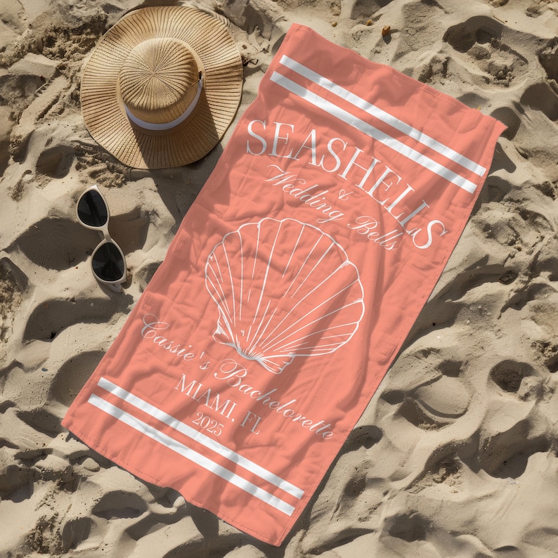 May include: Coral pink beach towel with white text that reads "Seashells & Wedding Bells" and "Cassie's Bachelorette Miami, FL 2023". A white seashell graphic is in the centre of the towel.