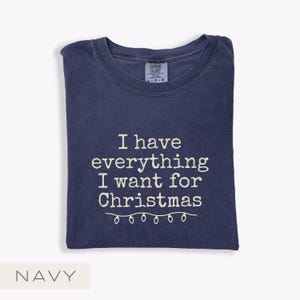 May include: Navy blue t-shirt featuring the white text "I have everything I want for Christmas" and a string of Christmas lights. The shirt is folded. The Comfort Colors tag is visible.