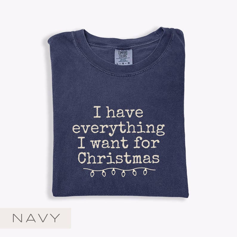 May include: Navy blue t-shirt featuring the white text "I have everything I want for Christmas" and a string of Christmas lights. The shirt is folded. The Comfort Colors tag is visible.