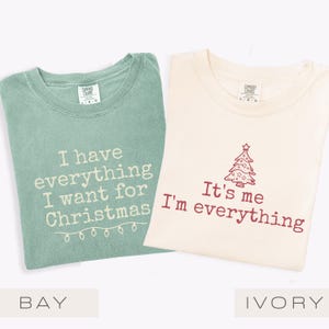 May include: Two t-shirts, one in bay green and one in ivory, with festive text. The bay green shirt reads "I have everything I want for Christmas" with fairy lights. The ivory shirt says "It's me I'm everything" with a Christmas tree design.