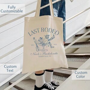 Op de afbeelding: Een beige tote bag met een blauw ontwerp met de tekst "Last Rodeo" met een vrouw in een cowboyhoed die in een martini-glas zit. De tekst zegt ook "Est. 2025" en "Nicole's Bachelorette Nashville, TN".