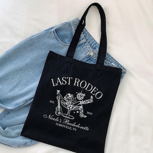Op de afbeelding: Zwarte tote bag met witte tekst die "Last Rodeo Est. 2025 Nicole's Bachelorette Nashville, TN" luidt. De tas heeft een witte illustratie van een vrouw met een cowboyhoed die een drankje en een gitaar vasthoudt.