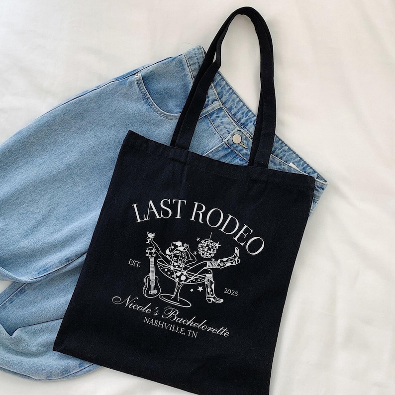 Op de afbeelding: Zwarte tote bag met witte tekst die "Last Rodeo Est. 2025 Nicole's Bachelorette Nashville, TN" luidt. De tas heeft een witte illustratie van een vrouw met een cowboyhoed die een drankje en een gitaar vasthoudt.