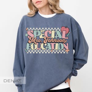 Op de afbeelding: Een denim blauwe sweatshirt met de tekst "SPECIAL Mrs. Johnson EDUCATION" in kleurrijke retroletters. Het ontwerp bevat een appel en sterren, met een geruite achtergrond. Het woord "DENIM" staat linksonder.