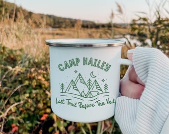 May include: White enamel camping mug with a green design of mountains, a campfire, a crescent moon, and stars. The text "Camp Hailey Last Trail Before The Veil" is printed on the mug.