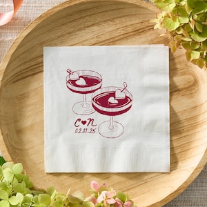 Può includere: Tovagliolo da cocktail di carta bianca con un'illustrazione rossa di due bicchieri di vino con cuori in cima. Il testo "C ♥ N 02.01.25" è stampato sotto i bicchieri.