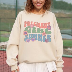 Puede incluir: Una sudadera beige con el texto "Pregnant Girl Summer" en una fuente retro con colores rosa, verde y azul. El texto est&aacute; rodeado de peque&ntilde;as flores rosas, moradas y azules.