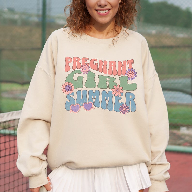 Puede incluir: Una sudadera beige con el texto "Pregnant Girl Summer" en una fuente retro con colores rosa, verde y azul. El texto est&aacute; rodeado de peque&ntilde;as flores rosas, moradas y azules.