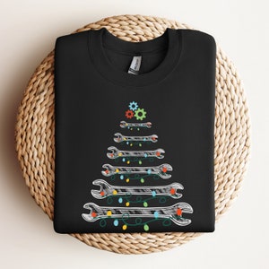 Puede incluir: Sudadera negra con un diseño de árbol de Navidad de llave inglesa blanca. El árbol está decorado con luces de colores y engranajes.