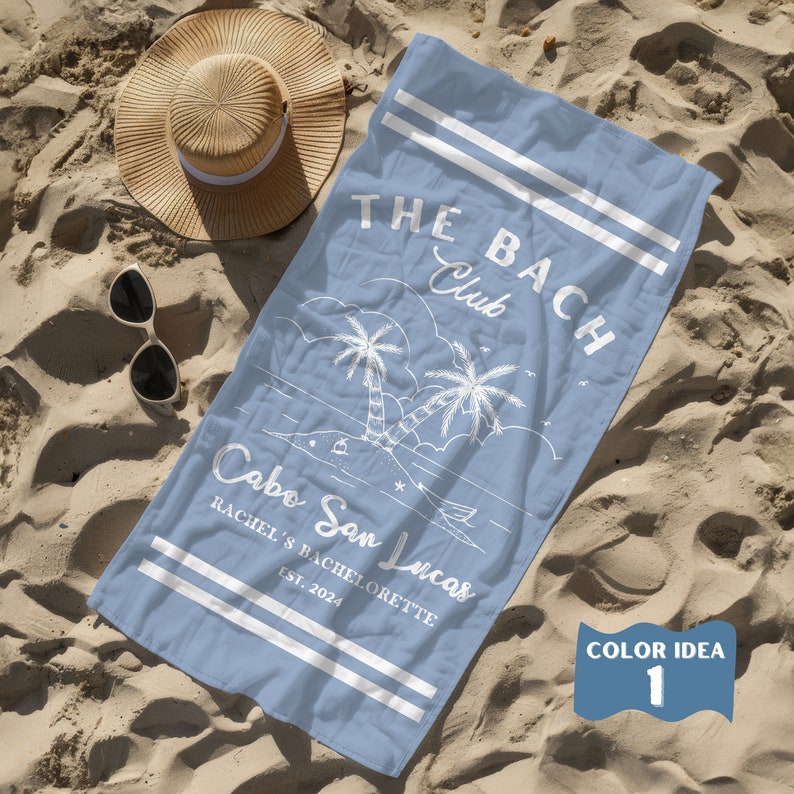 May include: A light blue beach towel with white text that reads "THE BACH Club Cabo San Lucas Rachel's Bachelorette Est. 2024". The towel features a white illustration of palm trees and a boat.