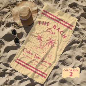 May include: A yellow beach towel with pink text that reads "THE BACH Club Cabo San Lucas Rachel's Bachelorette Est. 2024". The towel features a pink illustration of palm trees and a beach scene. The text "COLOR IDEA 2" is written in pink on a yellow circle in the bottom right corner of the towel.