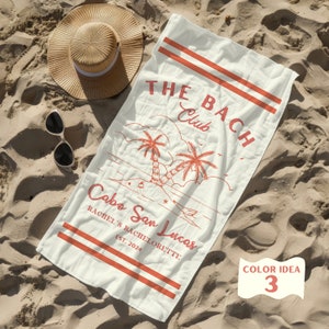 May include: A white beach towel with red text and graphics. The text reads "THE BACH Club Cabo San Lucas Rachel's Bachelorette Est. 2024". The graphic is of two palm trees on a beach.