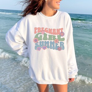 Puede incluir: Sudadera blanca con un gr&aacute;fico de estilo retro que dice "Pregnant Girl Summer" en letras de colores con flores y gafas de sol en forma de coraz&oacute;n.