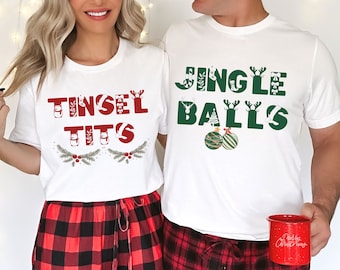 May include: A couple wearing matching white t-shirts with Christmas-themed text. The woman's shirt says "Tinsel Tits" and the man's shirt says "Jingle Balls". Both shirts feature green and red festive decorations.
