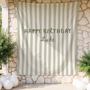 Puede incluir: Una pancarta de tela a rayas verticales verdes y blancas con el texto "HAPPY BIRTHDAY Luke". La pancarta est&aacute; colgada al aire libre, con vegetaci&oacute;n y globos blancos en primer plano.