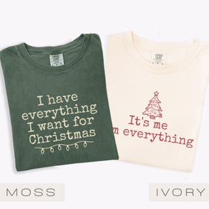 May include: Two t-shirts, one moss green and one ivory, with Christmas-themed text. The moss green shirt says "I have everything I want for Christmas" and the ivory shirt says "It's me everything" with a Christmas tree graphic.