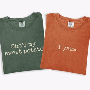 以下が含まれることがあります： 緑とオレンジの2枚のTシャツが表示されています。緑のシャツには白い文字で「She's my sweet potato」と書かれています。オレンジ色のシャツには小さなハートマークとともに「I yam」と書かれています。どちらも半袖です。