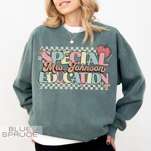 Op de afbeelding: Een blauwgroene sweatshirt met de tekst "SPECIAL Mrs. Johnson EDUCATION" in een kleurrijk retro-lettertype. Het ontwerp bevat een appel, sterren en een geruite achtergrond. De sweatshirt is een comfortabele, casual stijl.