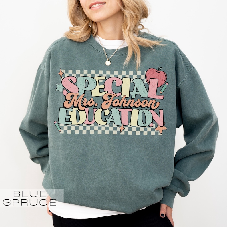 Op de afbeelding: Een blauwgroene sweatshirt met de tekst "SPECIAL Mrs. Johnson EDUCATION" in een kleurrijk retro-lettertype. Het ontwerp bevat een appel, sterren en een geruite achtergrond. De sweatshirt is een comfortabele, casual stijl.