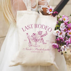 Op de afbeelding: Een witte canvas tote bag met een roze ontwerp met de tekst "Last Rodeo" met een afbeelding van een vrouw in een cowboyhoed die in een champagne glas zit met een gitaar en een discobal. De tekst onder de afbeelding zegt "Est. 2025 Nicole's Bachelorette Nashville, TN."