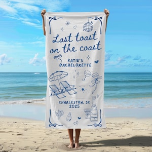 May include: White beach towel with blue text that reads "Last toast on the coast Katie's Bachelorette Charleston, SC 2025". The towel also features a blue illustration of a beach umbrella, two lounge chairs, champagne glasses, and a bottle of champagne.